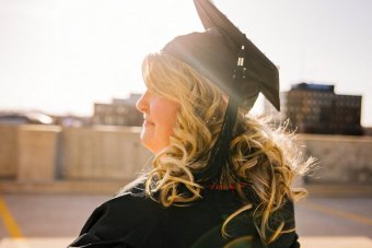 Photo credit: Esther Tuttle. A white woman, in profile, stands wearing a black graduation cap and gown