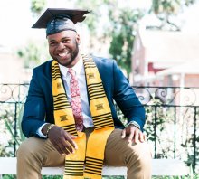 Photo credit: William Stitt. A young black man in a graduation cap and gown sits smiling on a bench in a garden