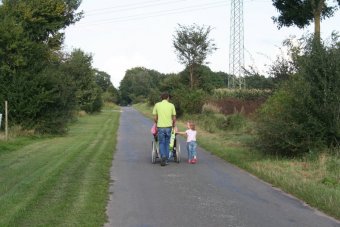 A photo of a family, taken from the back. A man in a green shirt pushed someone using a wheelchair, while walking with a small child.